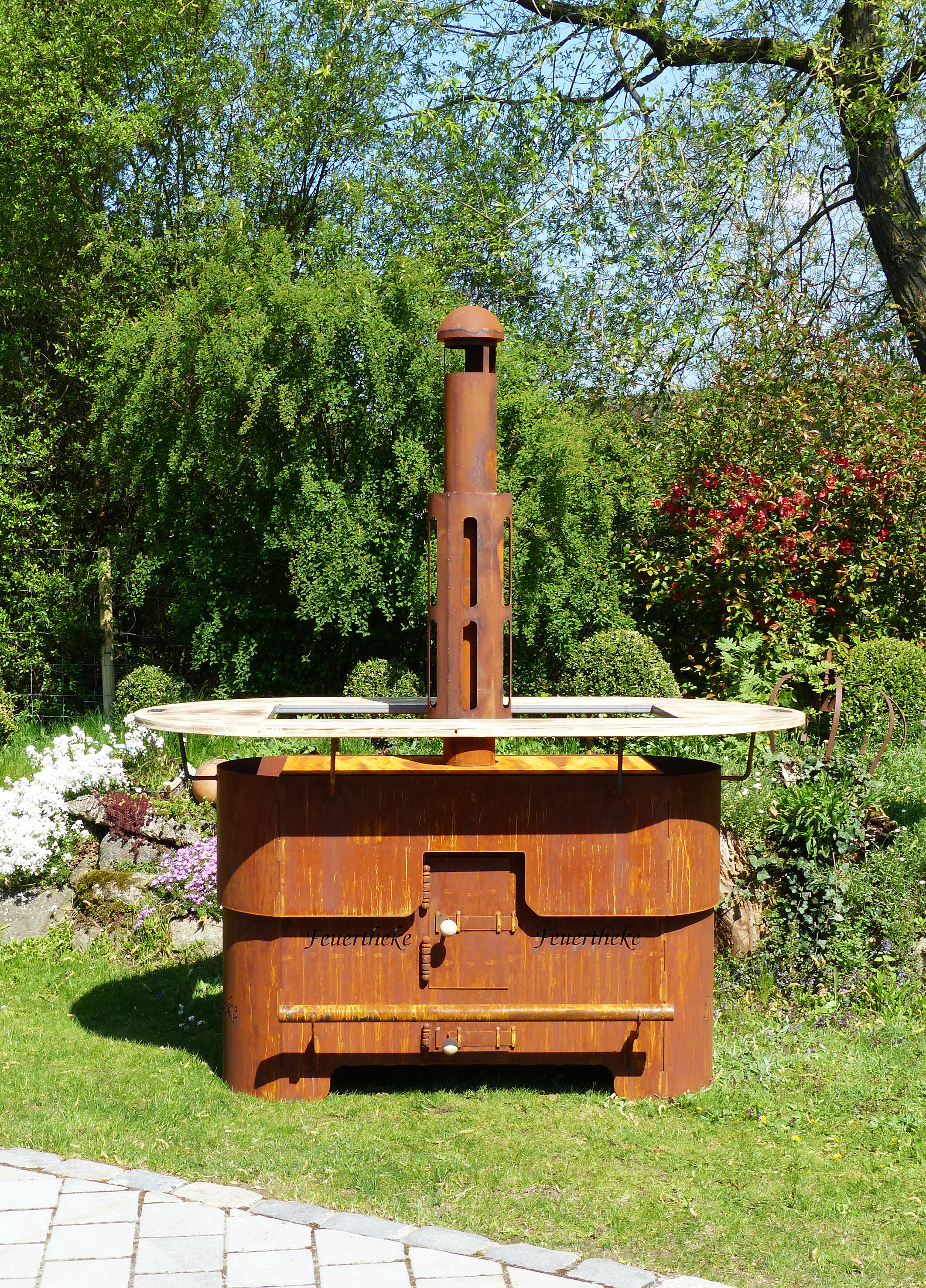 Feuertheke aus Edelrost mit Holzablage im Garten als dekorative Feuerstelle im Außenbereich