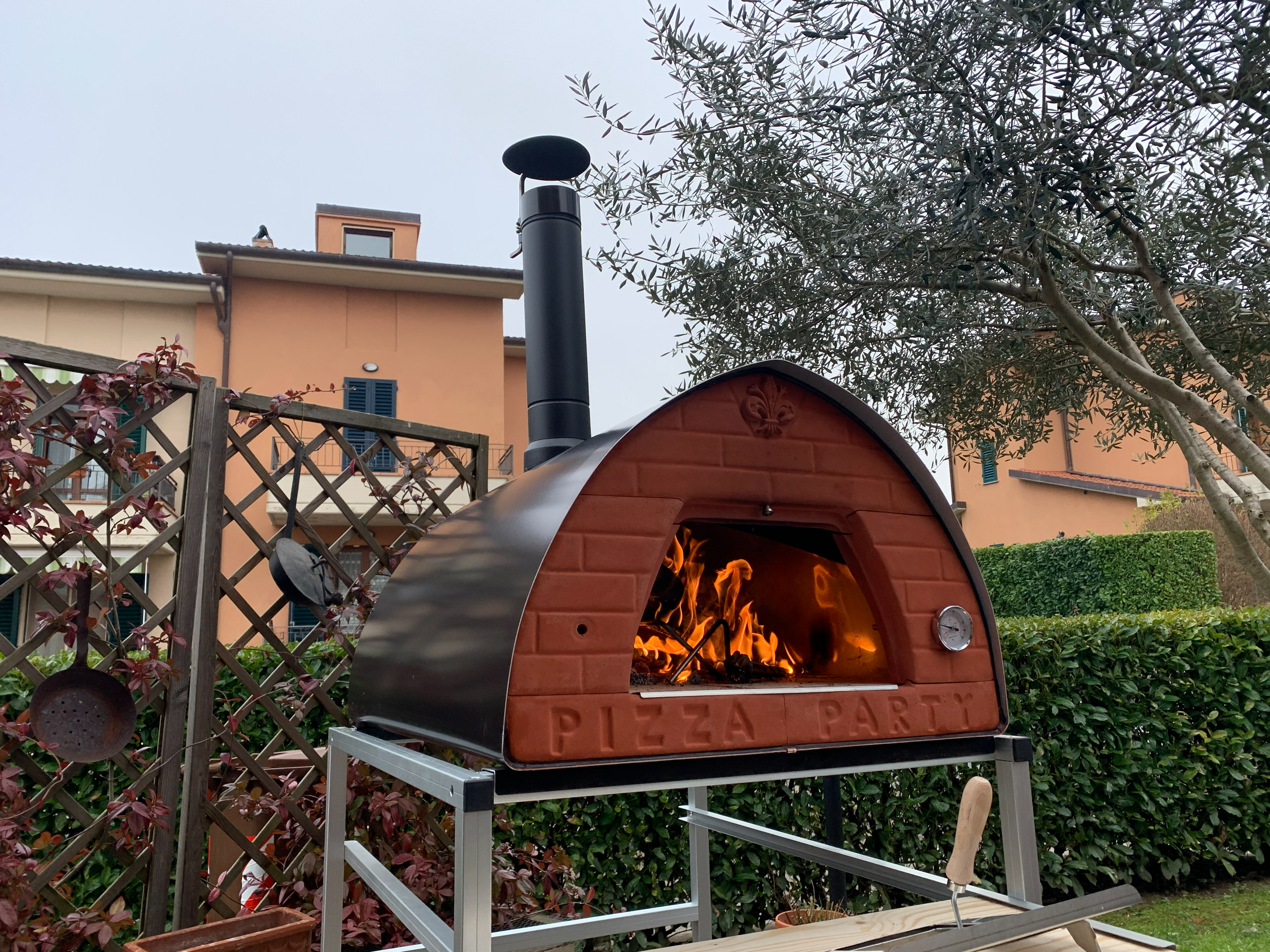 Befeuerter PizzaParty 70x70 Holzbackofen auf silberfarbenem Rollgestell bei der Verwendung im Garten.