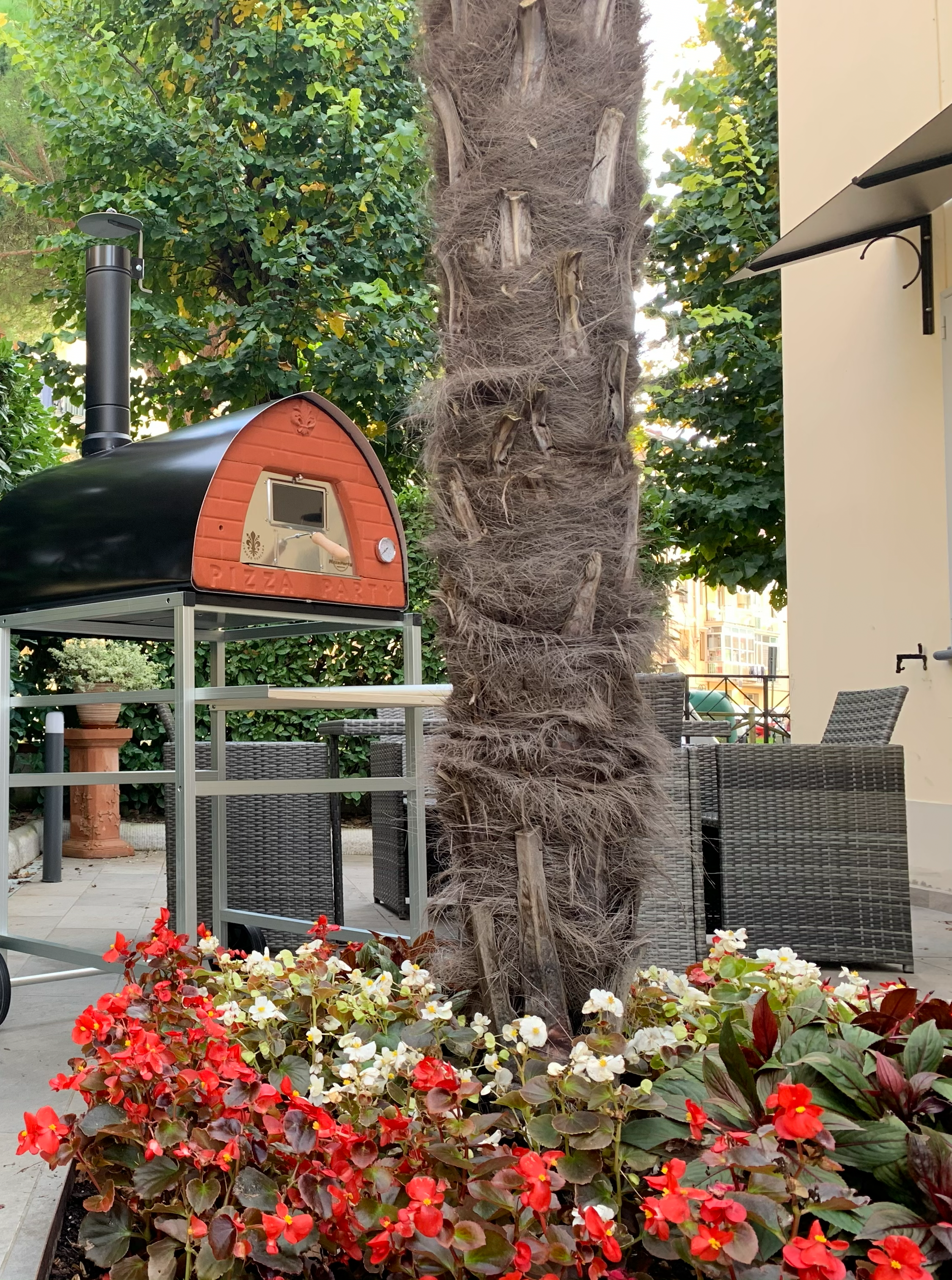 PizzaParty 70x70 Gunmetal mit Kamin bei der Aufstellung im Garten zwischen Bäumen und blühenden Pflanzen.