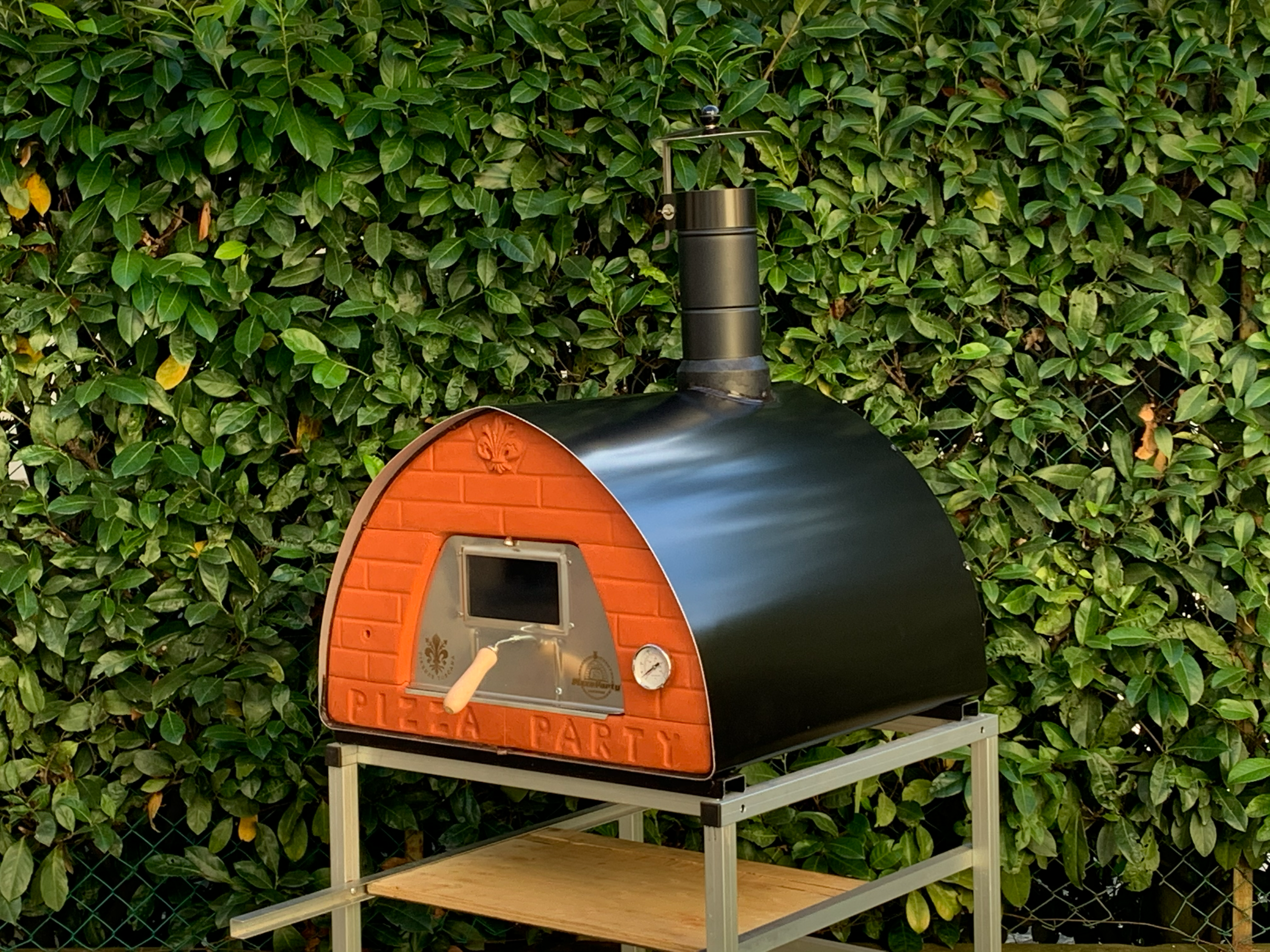 PizzaParty 70x70 Holzbackofen mit Terrakottafront und Gunmetal-Gehäuse vor einer grünen Gartenhecke.