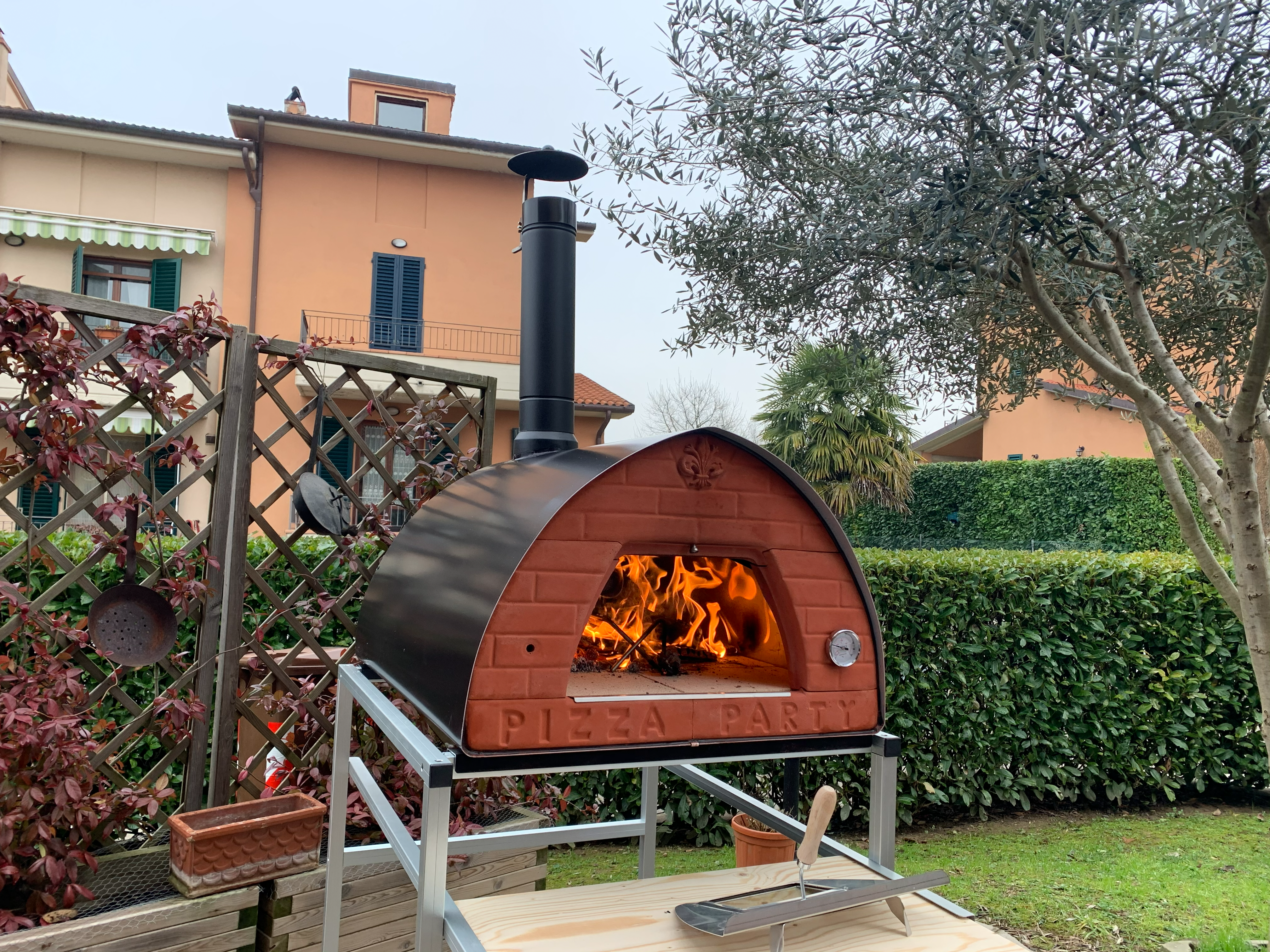 PizzaParty 70x70 Gunmetal auf einem mobilen silberfarbenen Untergestell mit Holzablage während des Betriebs im Garten.